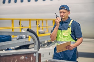 Front view of a serious airport worker with a walkie-talkie standing near the conveyor belt