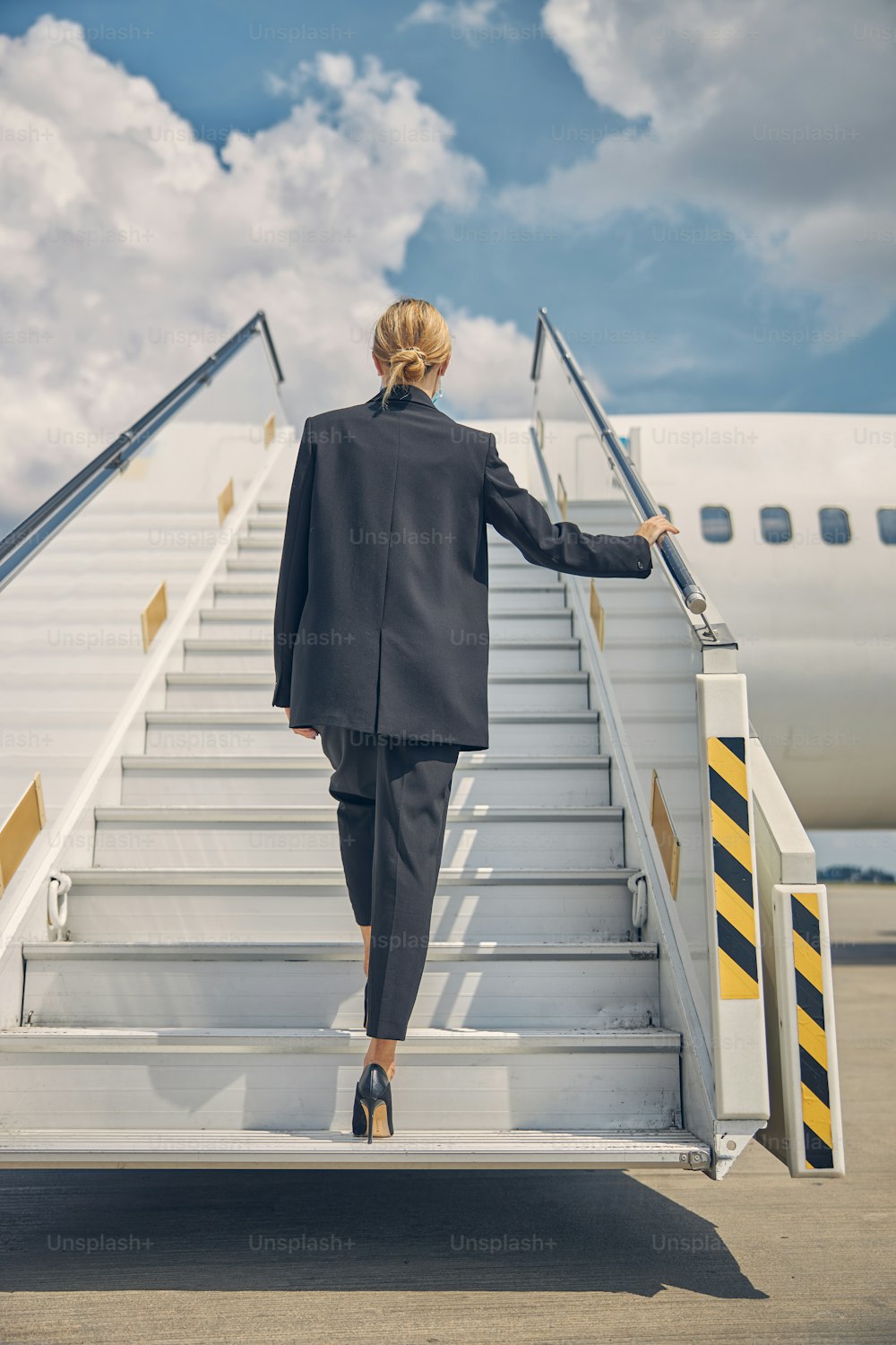 Vista trasera de una delgada dama rubia con un traje de pantalón subiendo los escalones del avión