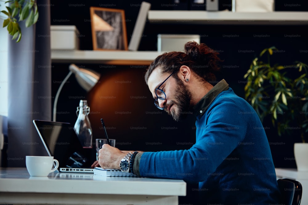 Side view of young attractive bearded freelancer sitting in his home office and writing tasks in notebook.