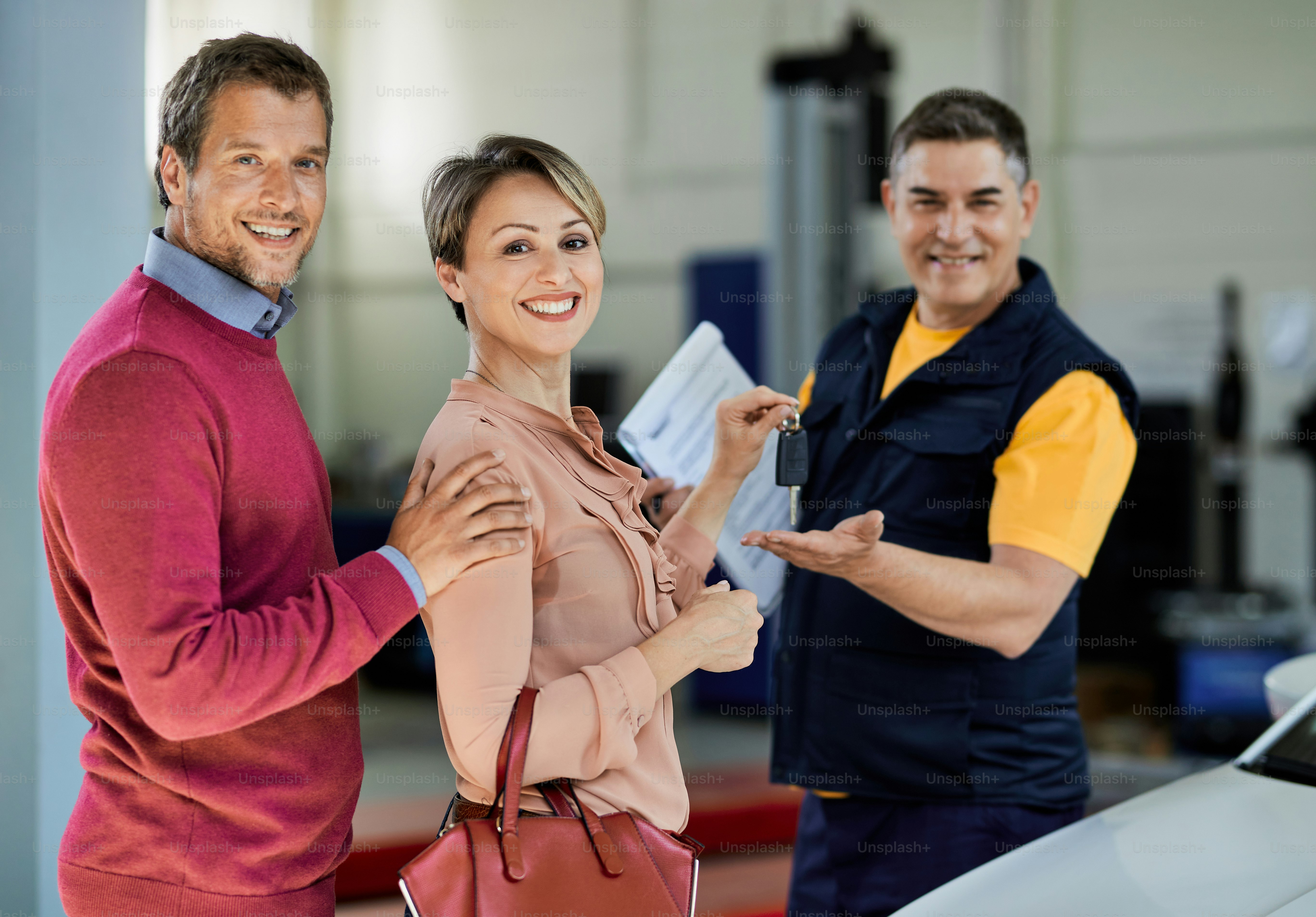 Happy couple giving car key to their auto repairman while looking at camera in a repair shop.