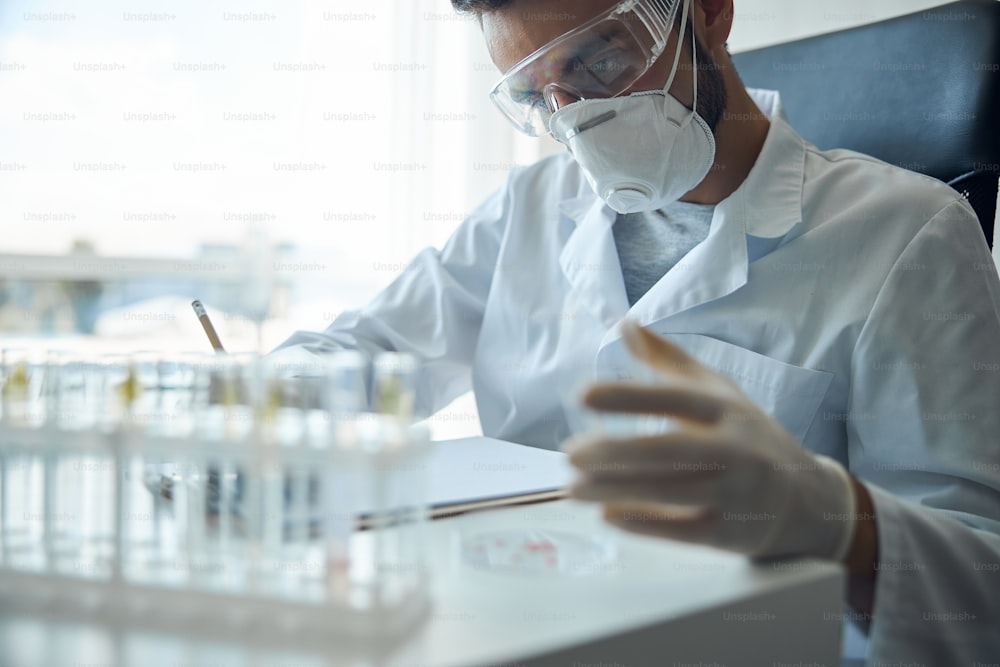 Cientista do sexo masculino focado em óculos de segurança e uma máscara facial trabalhando com uma amostra de sangue