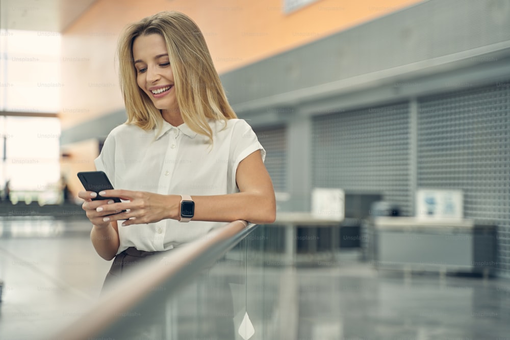 Erfreute junge Frau, die ein Lächeln auf ihrem Gesicht behält, während sie auf den Bildschirm ihres Smartphones schaut