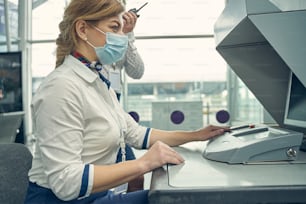 Alegre mulher de cabelos compridos usando máscara médica enquanto trabalha no aeroporto com passageiros