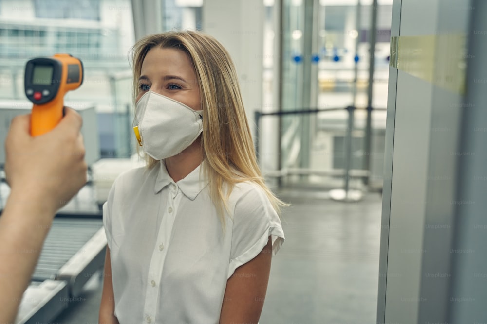 Pretty female wearing protective mask while standing opposite the worker of airport and checking temperature