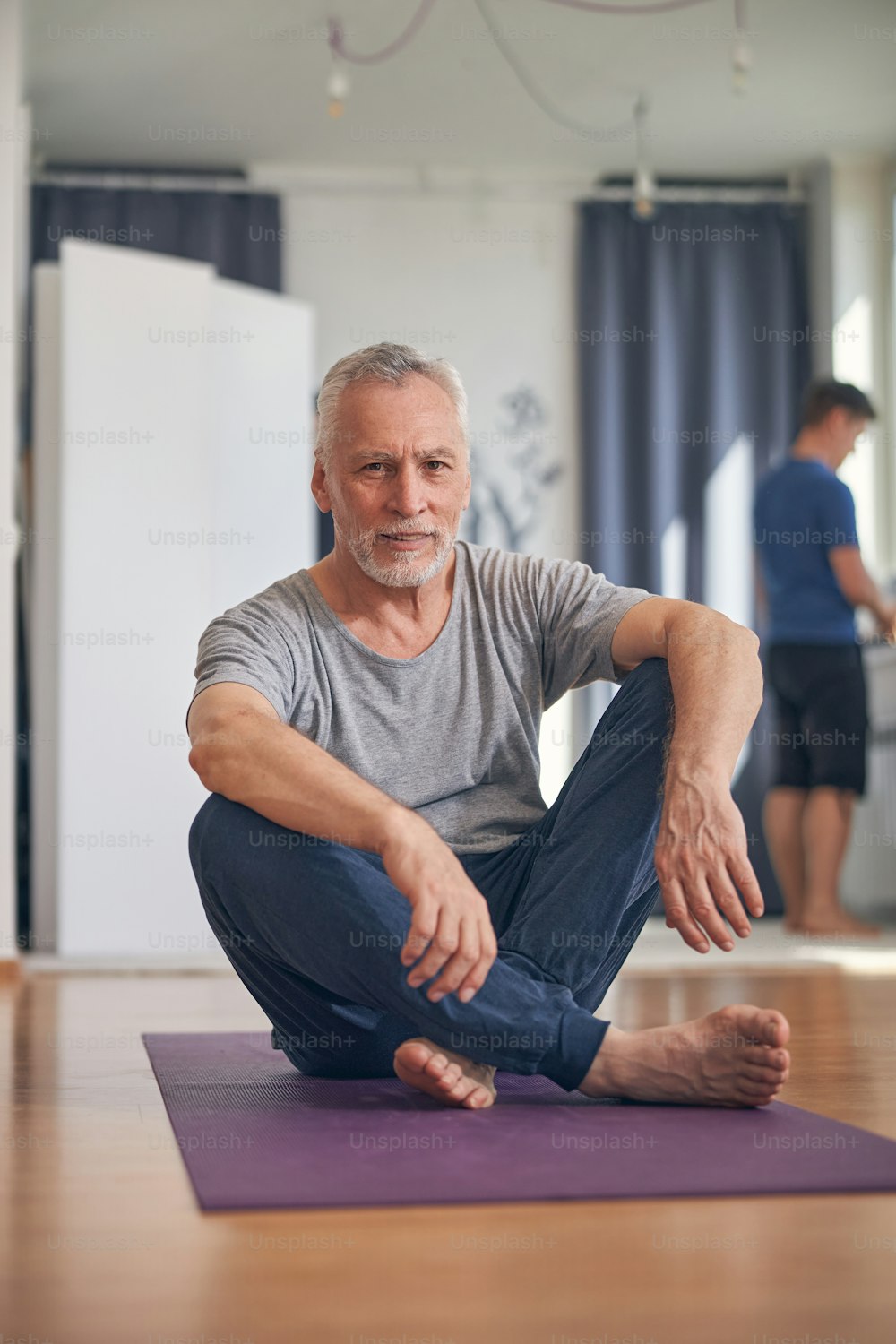 Front view of a grey-haired male sitting on a yoga mat in a fitness studio