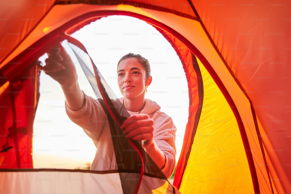 Attraente viaggiatrice femminile in felpa che apre la cerniera della tenda turistica e sorride
