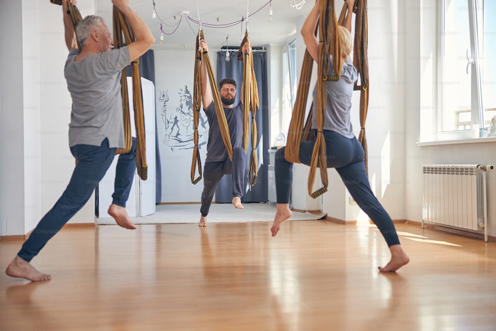 Serene young Caucasian professional trainer performing an aerial warrior pose two with two beginner yogis