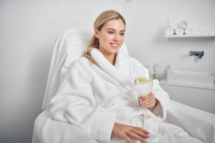 La mujer hermosa y alegre está sentada en una silla de pedicura en bata de baño y disfruta de una bebida refrescante con limón
