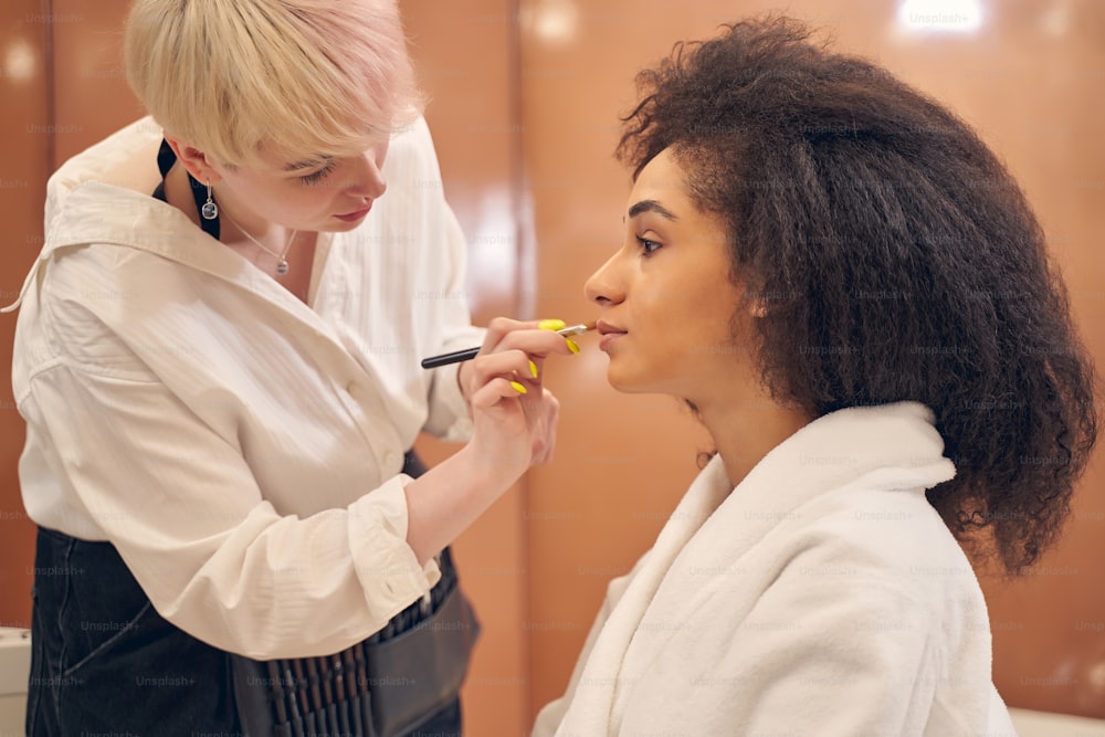 Concentrated makeup artist using a brush and applying concealer while adorable curly woman sitting calmly