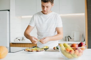 Home cooking concept. Man putting avocado slices on sandwich made of whole grain bread,  tomato, and cheese. Process of making toasts with avocado on modern white kitchen.