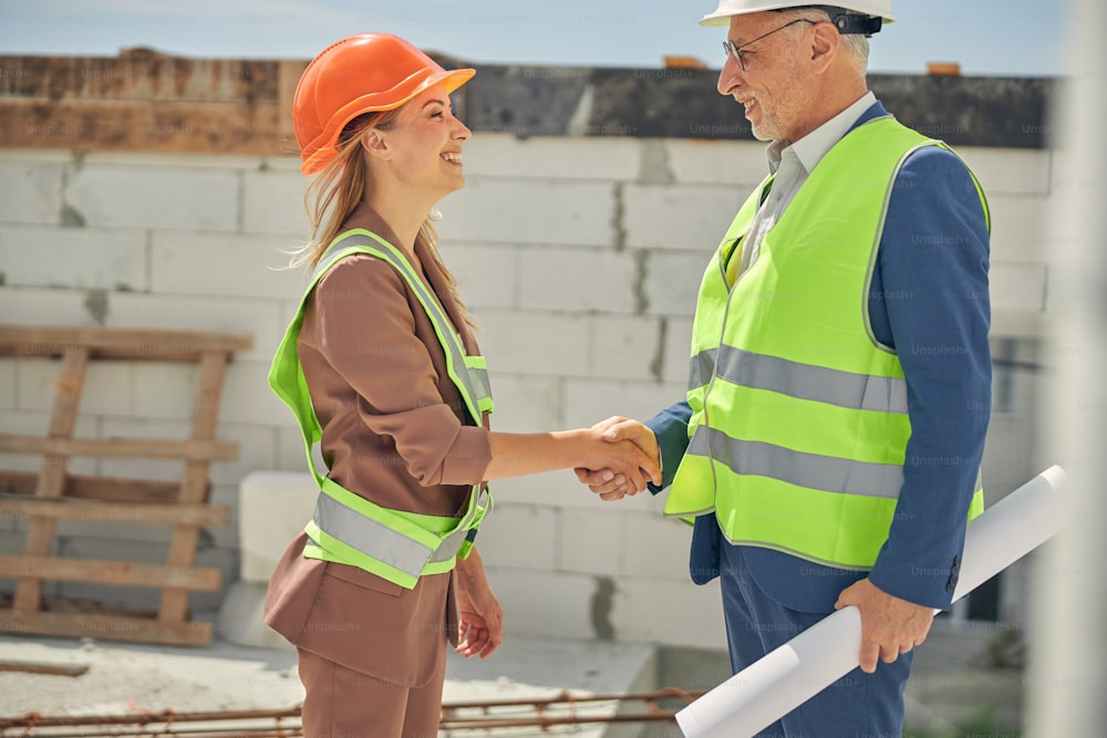 Side view of a merry female and a male constructor greeting each other at the building site