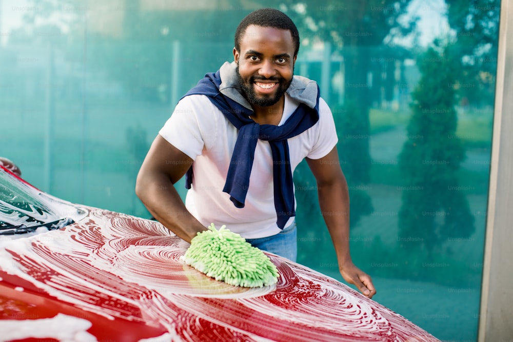Car washing, outdoor self car wash service concept. Smiling African young man cleaning his red car using sponge and foam, looking at camera.
