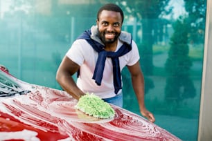 Car washing, outdoor self car wash service concept. Smiling African young man cleaning his red car using sponge and foam, looking at camera.