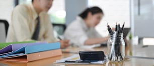 A calculator is putting on the wooden table surrounded by various office equipment over employees as a background.