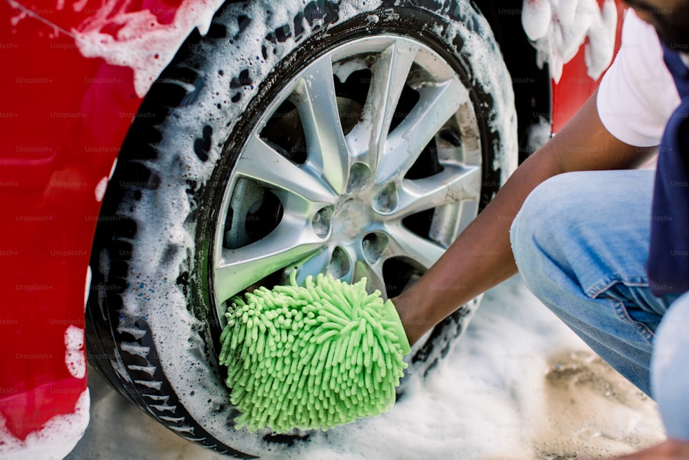 Hände eines afrikanischen Mannes, der grünen Schwamm hält und Autorad mit Schaum wäscht. Reinigung von modernen Felgen des roten Luxusautos bei Selbstwaschanlage im Freien.