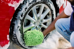 Mani dell'uomo africano che tiene la spugna verde, lavando la ruota dell'automobile con la schiuma. Pulizia dei moderni cerchioni di auto rosse di lusso presso il servizio di autolavaggio all'aperto.
