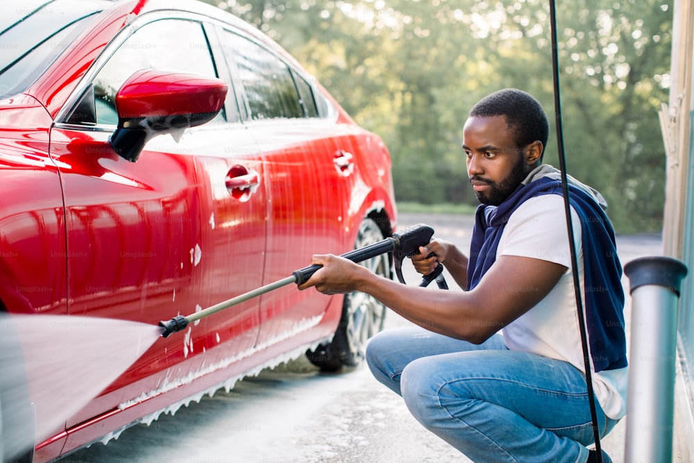 Jeune homme africain en tenue décontractée lavant sa voiture de luxe à la station de lavage de voiture à l’extérieur, en utilisant un jet d’eau à haute pression