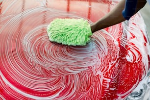 Nahaufnahme abgeschnittenes Bild der Hand eines afrikanischen Mannes mit grünem Schwamm, der seine rote Motorhaube in einer Selbstbedienungswaschanlage im Freien wäscht. Motorhaube des roten Luxusautos mit Seife bedeckt