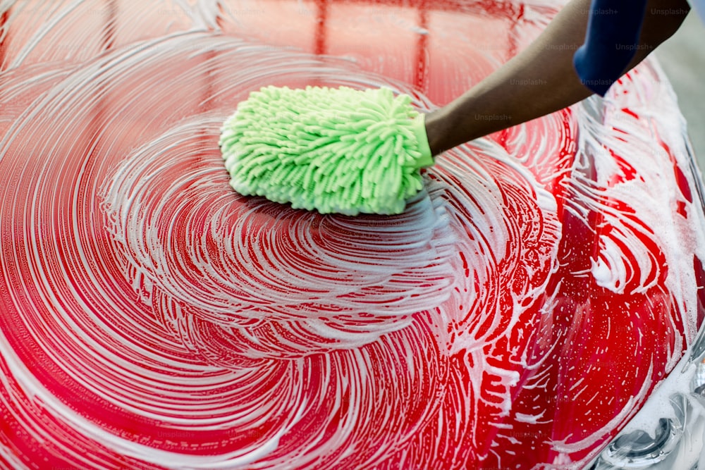 Image recadrée en gros plan de la main d’un homme africain avec une éponge verte lavant son capot de voiture rouge dans un lave-auto en libre-service à l’extérieur. Capot d’une voiture de luxe rouge recouvert de savon