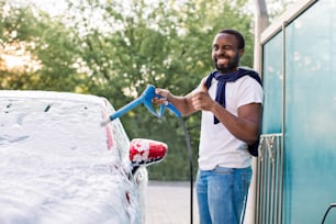 Un uomo afroamericano ammiccante felice, all'autolavaggio all'aperto, sta spruzzando schiuma detergente su una moderna auto di lusso rossa che tiene un'alta pressione e mostra il pollice in su.