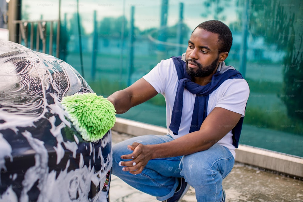 Car washing outdoors ar self wash service station. Handsome African dark skinned man in casual wear, washing his modern electric car headlights with green sponge and soap.