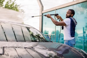 Handsome African young man in casual wear, washing his red car manually with water high-pressure hose at outdoor self wash service. Car washing concept.