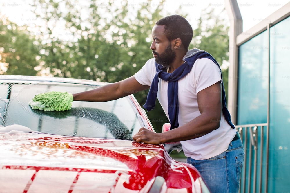 Handsome African man cleaning his red car windshield with green sponge and soap foam outdoors at car wash service.