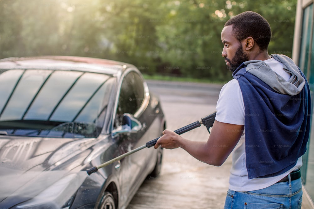 Servicio de lavado de autos al aire libre. Vista lateral de un chico africano con camiseta blanca y jeans, lavando un automóvil de lujo eléctrico azul con pistola de agua en un lavado de autos al aire libre