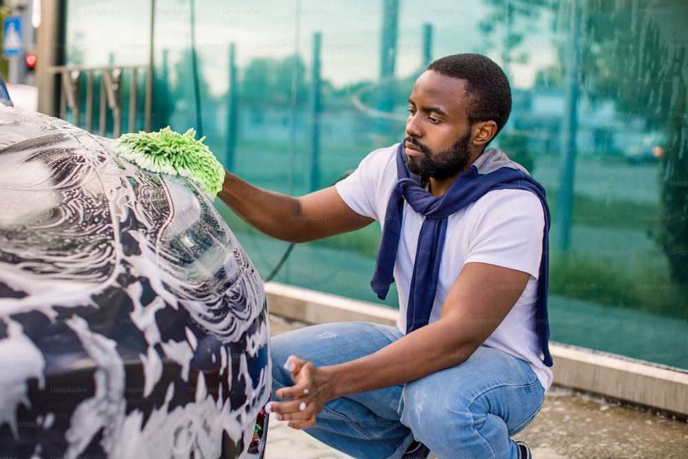 Joven africano guapo y barbudo en camiseta y jeans limpiando su lujoso automóvil gris eléctrico al aire libre en la estación de lavado de autos, usando trapo verde y espuma de jabón