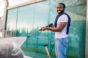 Manual car wash. Handsome African young man washing his luxury vehicle with high pressure water pump at automobile cleaning self service outdoors