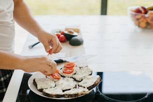 Pessoa colocando fatia de tomate em creme de queijo em pão integral na cozinha branca moderna. Processo de fazer torradas saudáveis com abacate, tomate, rúcula e queijo. Conceito de culinária caseira