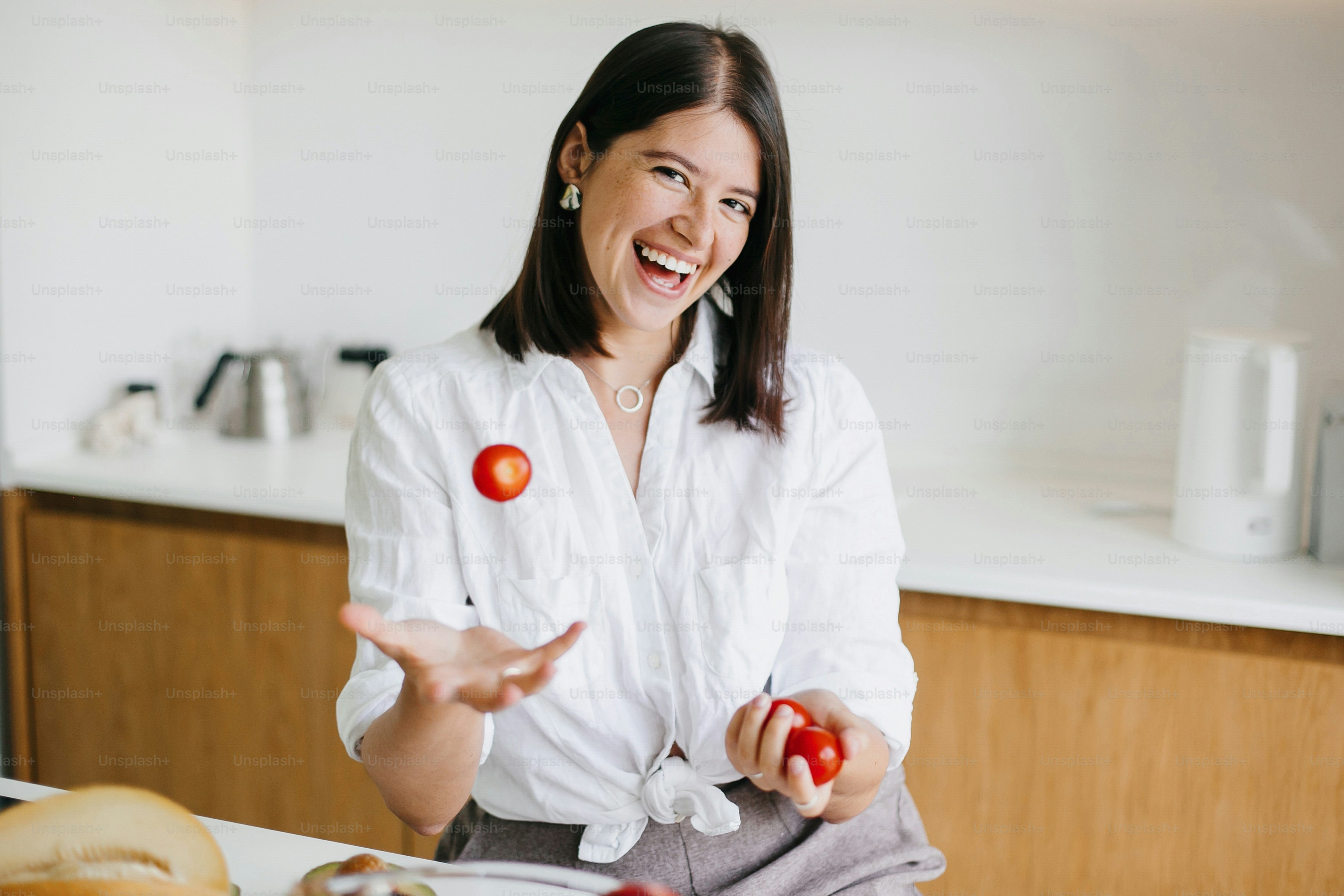 チェリートマトを投げ、モダンな白いキッチンの背景に微笑む若い幸せな女性。健康食品のコンセプト。家庭料理、サラダの具材