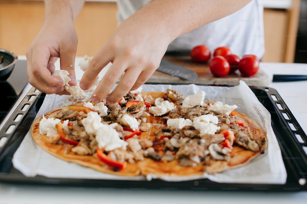 Hausgemachte Pizza. Person, die Mozzarella auf Teig mit Ketchup, Huhn und Pilzen in der modernen weißen Küche aufträgt. Prozess der Herstellung von Pizza, Zutaten aus nächster Nähe. Hausmannskost-Konzept