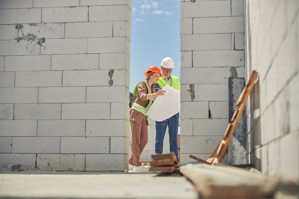 Smiling blonde lady and a man in protective helmets and reflective vests staring at house plans