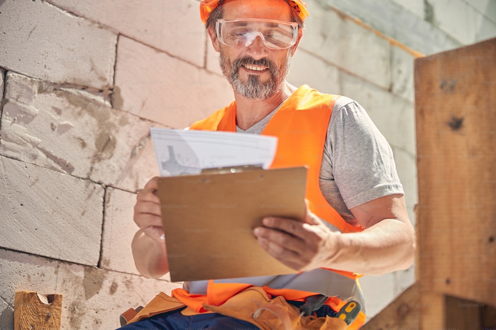 Lächelnder fröhlicher Bauingenieur mit Schutzbrille schaut auf einen Hausplan in seinen Händen