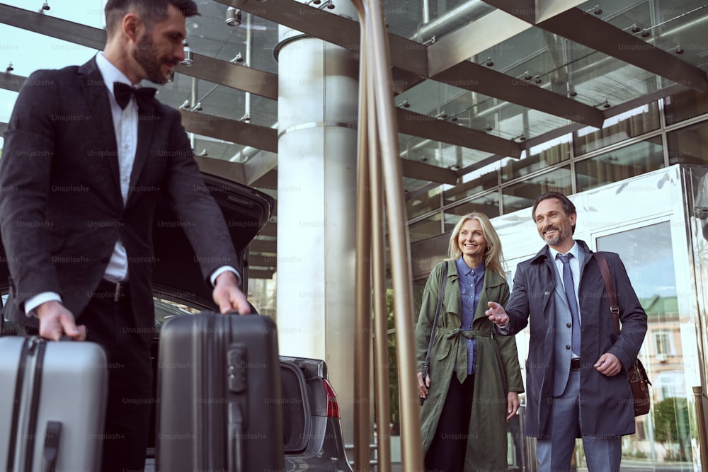 ホテルの近くに立っている陽気な男と笑顔の妻が、ポーターがスーツケースを扱ってくれるのを待っている
