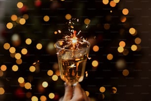 Happy New Year. Burning firework in champagne glass in hand on background of golden bokeh lights in festive room. Hand holding drink with sparkler at christmas tree. Creative aesthetic moment