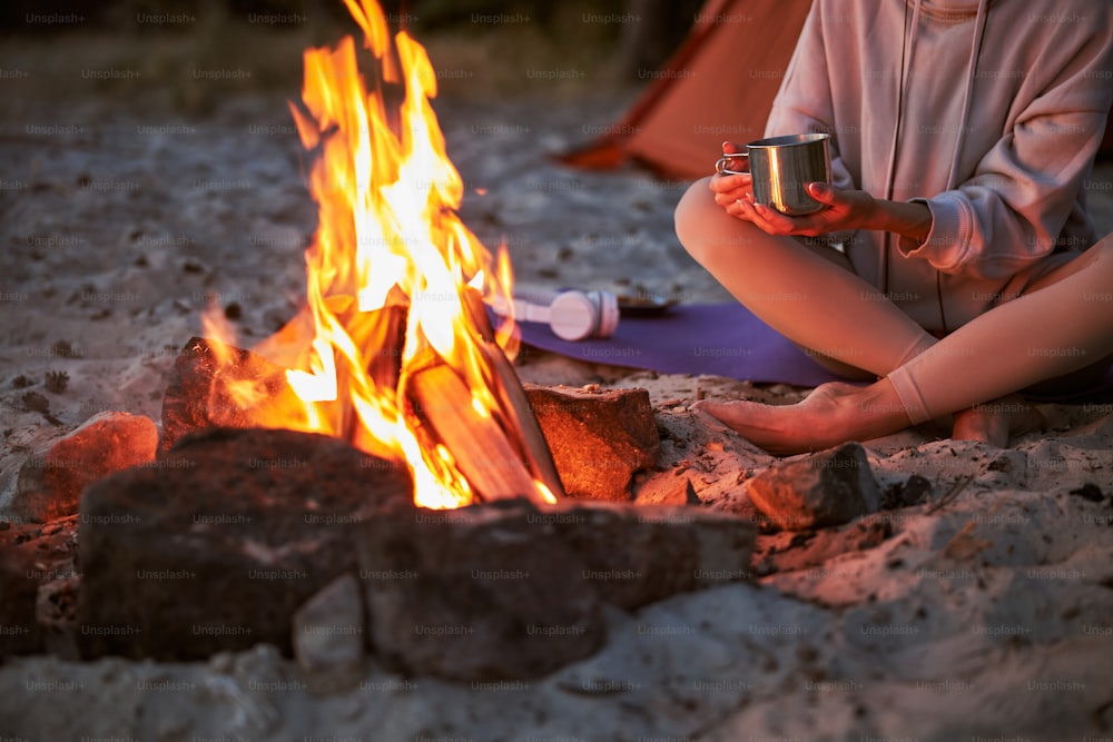 Close up of female traveler holding mug of hot drink while sitting on sand near bonfire