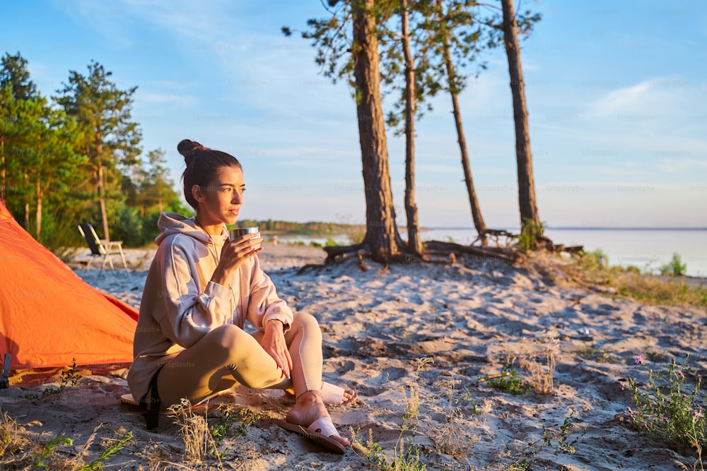 スウェットシャツを着た魅力的な女性旅行者が温かい飲み物のカップを持ち、砂の上に座りながら海の景色を楽しんでいます