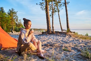 スウェットシャツを着た魅力的な女性旅行者が温かい飲み物のカップを持ち、砂の上に座りながら海の景色を楽しんでいます