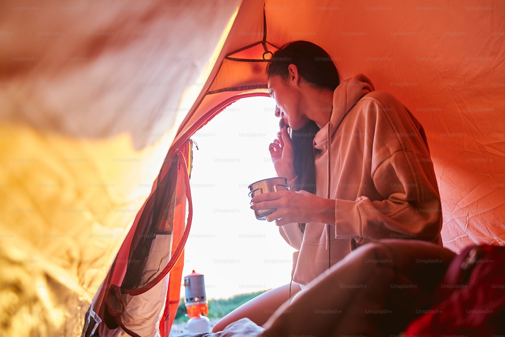 Charming female traveler holding mug of hot drink and eating cookie while sitting in touristic tent