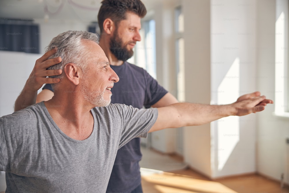 Grey-haired male doing a stretching yoga exercise assisted by a qualified instructor at a gym