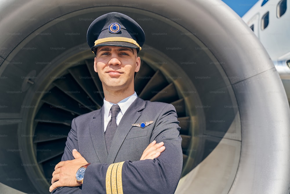 Retrato de un joven aviador sereno con los brazos cruzados de pie frente al motor de aviación