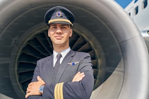 Porträt eines heiteren jungen Piloten mit verschränkten Armen, der vor dem Flugzeugtriebwerk steht
