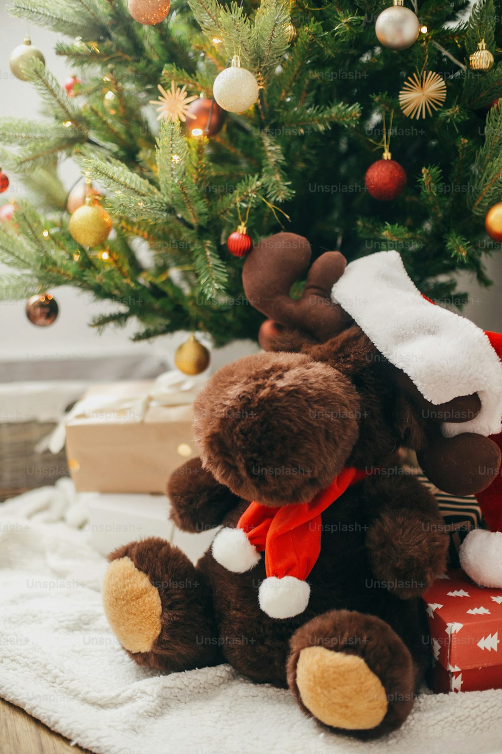 Lindo juguete de reno en sombrero de Papá Noel debajo del árbol de Navidad decorado moderno con regalos y luces festivas. Gran juguete sorpresa en el árbol de Navidad con adornos rojos y dorados, decoración de habitación escandinava