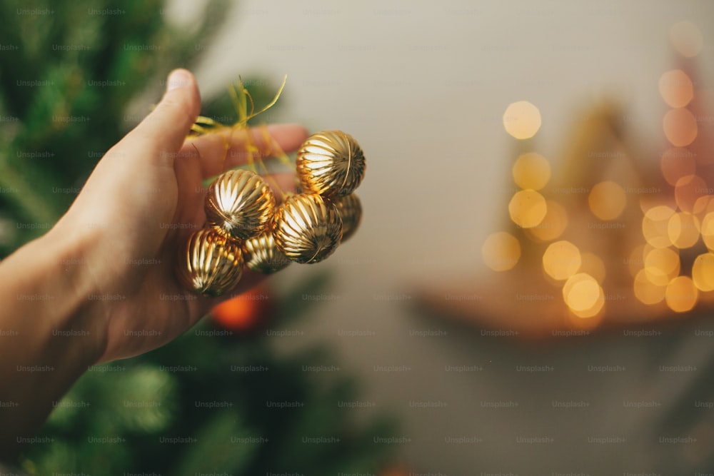 Hanging ornaments on christmas tree, festive decoration of room. Hand holding modern gold baubles on background of green tree. Decorating christmas tree.