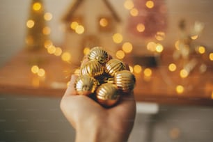 Mão segurando enfeites de ouro moderno no fundo luzes de iluminação desfocadas. Decorando árvore de Natal. Enfeites pendurados na árvore de Natal, decoração festiva da sala