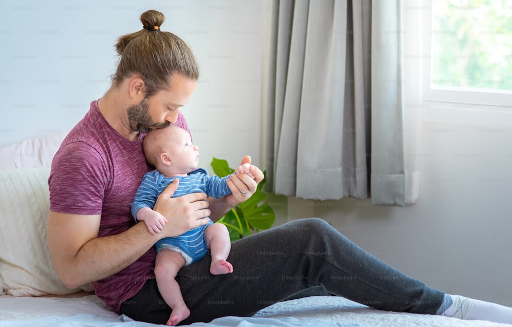 Caucasian father sitting on bed with holding and kissing his newborn baby son.