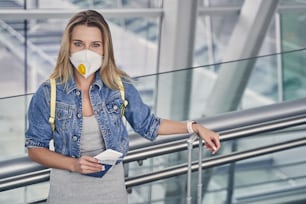 Attractive long haired woman wearing protective mask and holding boarding pass in right hand while waiting for flight