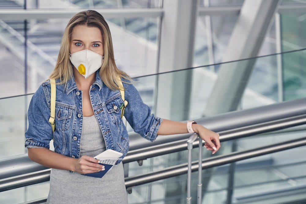 Attractive long haired woman wearing protective mask and holding boarding pass in right hand while waiting for flight